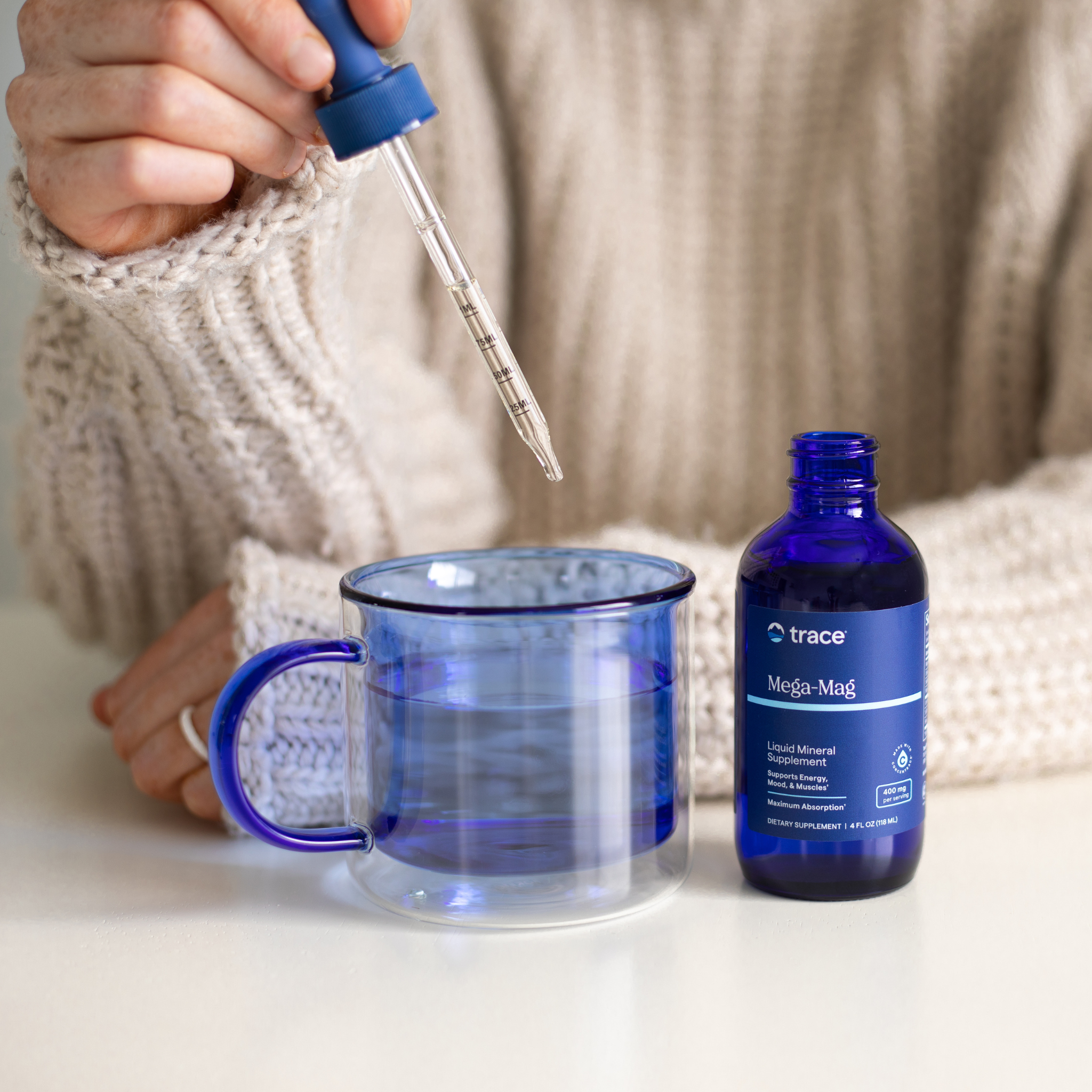 „Asmuo su megztiniu laiko pipetę virš mėlyno stiklinio puodelio su vandeniu, o šalia stovi atidarytas „Trace Mega-Mag“ buteliukas.