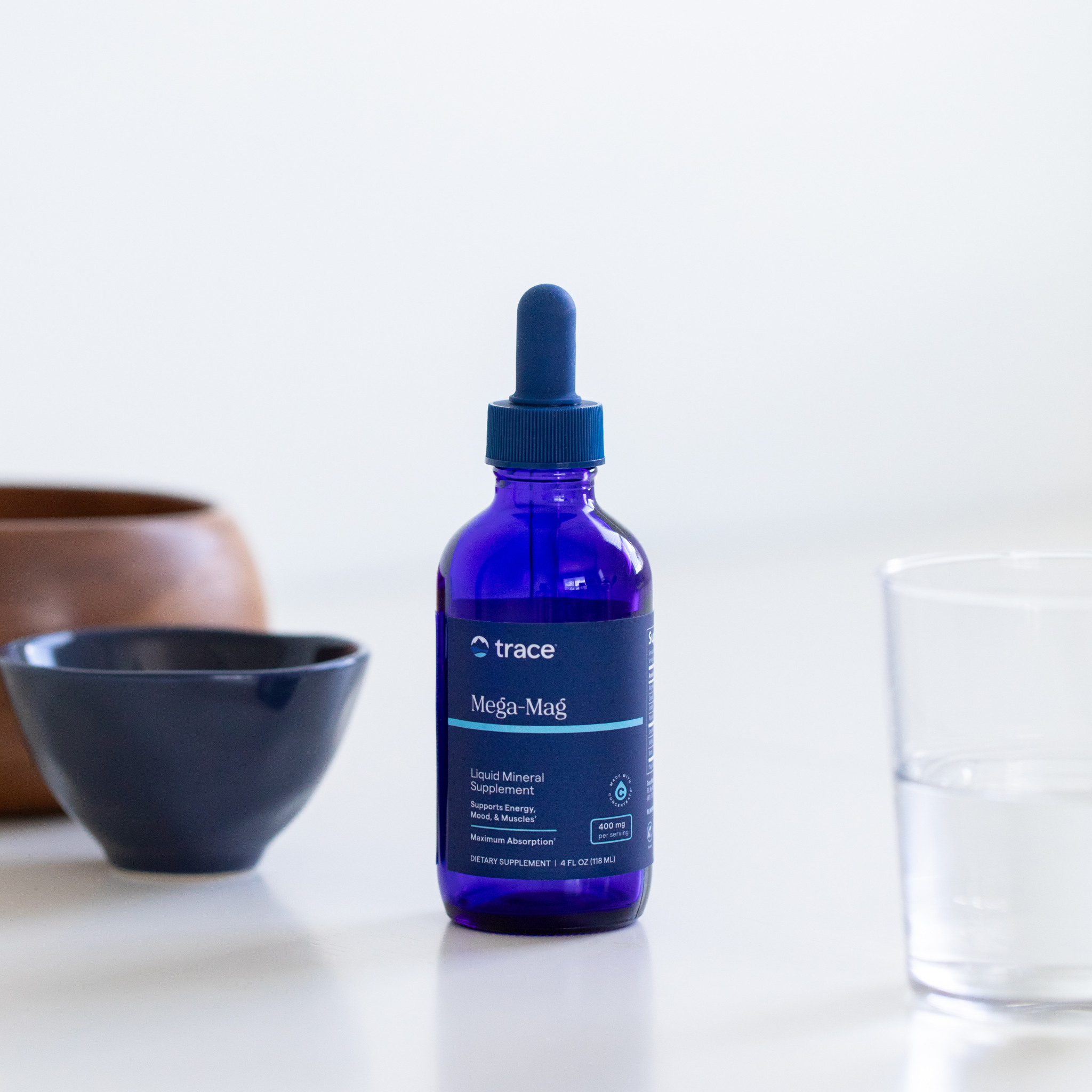 Mėlynas „Trace Mega-Mag“ skysčio buteliukas su pipete stovi ant šviesaus paviršiaus, šalia dubenėlio ir stiklinės vandens.